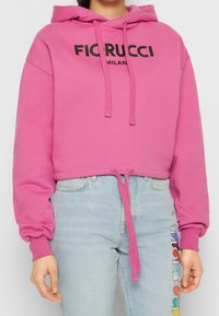 Sudadera rosa corta con capucha ajustable y letras negras que dicen "FICRUCCI MILAN". Combinada con vaqueros azul claro que llevan un logo colorido en el lateral.