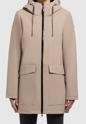 Beige-huppariparka, jossa musta etuvetoketju, säädettävät kiristysnauhat, pitkät hihat ja kaksi etutaskua, jota henkilö käyttää.