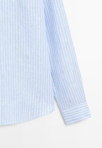 Chemise à manches longues bleu clair avec des rayures blanches verticales, dotée d'un poignet boutonné et d'un ourlet légèrement courbé sur fond blanc.
