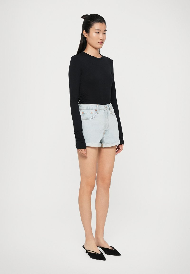 Zwarte lange mouwen top, lichtblauwe hoge taille spijkerbroek shorts en zwarte puntige ballerina's. Model staat met een ontspannen houding op een neutrale achtergrond.