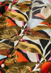 Tessuto con grande motivo di foglie autunnali in tonalità di marrone, giallo, arancio e rosa, con bottoni rivestiti in tessuto lungo il centro.