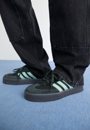 Stopy w czarnych i turkusowych butach sportowych Adidas oraz luźnych ciemnych dżinsach stojące na jasnoniebieskim dywanie.