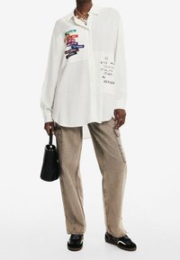 Camicia bianca oversize con bottoni, caratterizzata da etichette ricamate colorate, abbinata a pantaloni cargo beige e una borsa nera. Scarpe nere completano l'outfit.