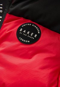 Część kurtki w kolorze czerwonym i czarnym, ocieplanej, z okrągłą czarną naszywką z białym napisem "BAKER by TED BAKER LONDON". Gładka tekstura, lekki materiał.
