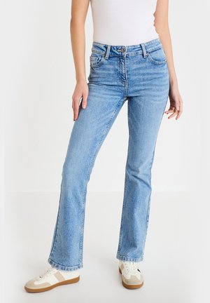 Osoba nosząca jasnoniebieskie dżinsy z wysokim stanem i prostymi nogawkami, białą bluzkę oraz beżowo-białe sneakersy, stojąca na jednolitym tle.