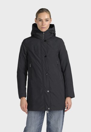 Femme portant un manteau d'hiver noir à capuche avec fermeture éclair et boutons à l'avant, debout devant un fond gris uni.