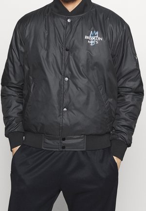 Schwarze Bomberjacke mit Druckknöpfen, die ein kleines Brooklyn Nets Logo auf der linken Brust sowie gerippte Bündchen und Saum hat.
