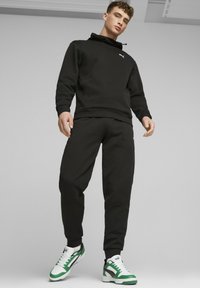 Sudadera con capucha negra y pantalones jogger a juego en una tela suave, con un logo blanco. El calzado incluye zapatillas verdes y blancas con acentos negros.
