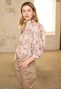 Pitkät vaaleatukkaiset nainen pukeutuu vaaleanpunaiseen paisley-kuvioiseen paitaan ja beigenvärisiin housuihin, seisoo käsineen taskuissa kirkkaassa huoneessa.