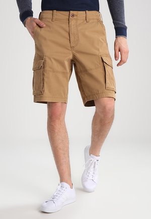 Khaki szorty cargo wykonane z trwałej tkaniny, z wieloma kieszeniami, prostym krojem i zapięciem na guzik. Noszone z białymi trampkami.