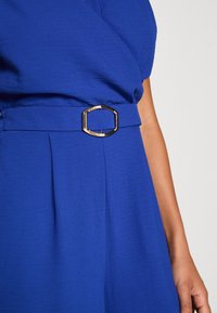 Blå klänning med en samlad tygstruktur och en bältesknäppning i guld med sexhörning. Plisserad detalj i midjan förbättrar designen.