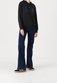 Felpa nera con logo "GAP" in strass, abbinata a jeans in denim scuro a zampa d'elefante e scarpe nere con suola chiara e strisce.