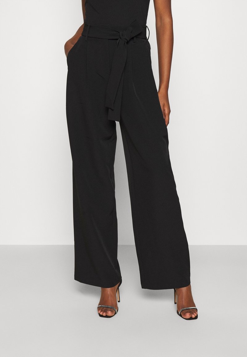 Femme portant un pantalon noir taille haute à jambes larges avec une ceinture en tissu nouée à la taille et des sandales noires à talons ouverts.