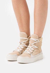 Sneakers beige alti con lacci bianchi, suole spesse bianche e rivestimento peloso indossati su gambe nude su sfondo bianco.