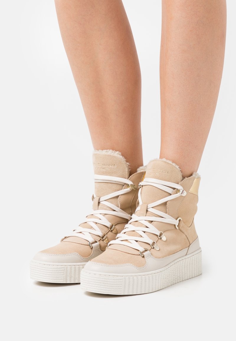 Sneakers beige alti con lacci bianchi, suole spesse bianche e rivestimento peloso indossati su gambe nude su sfondo bianco.
