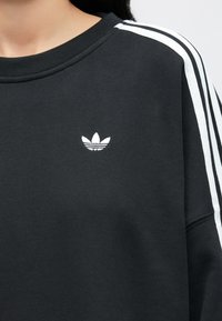 Čierna mikina s bielym vyšívaným logom na hrudi, s bielymi pruhmi na ramenách a rebrovaným výstrihom.