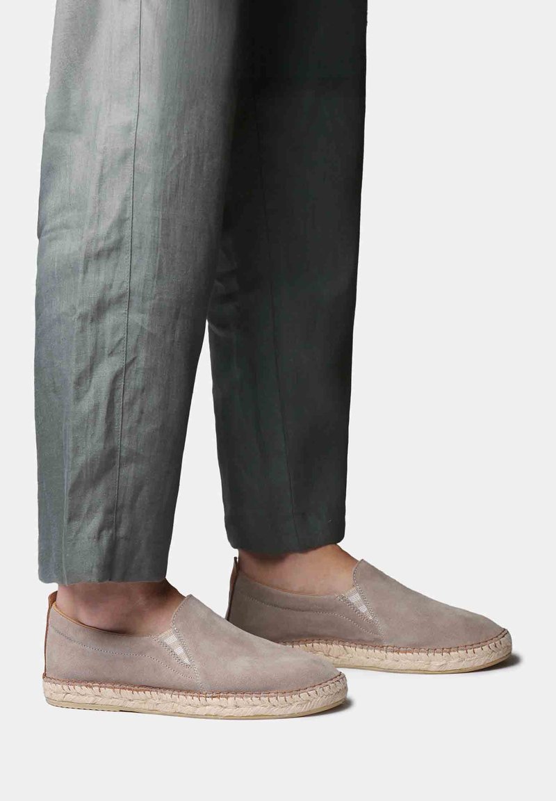 Jalad, mis kannavad heledat taupe'i värvi suss-laadsed espadrilles'id, millel on põimitud jootõlmetest põhjaga tallad, kombineeritud tumehallide lühendatud pükstega, taustal on lihtne voog.