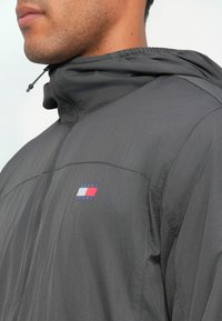 Szara lekka kurtka z kapturem, charakteryzująca się subtelną teksturą, zapinana na zamek z przodu oraz małym logo "TOMMY JEANS" z czerwonymi, białymi i niebieskimi akcentami.