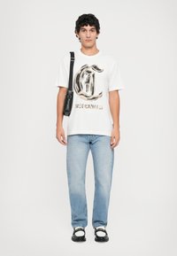 Hvit t-skjorte med svart og gull grafikk, blå denimjeans og svarte sko med hvite detaljer. Crossbody veske i svart lær.