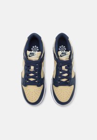 Paire de baskets Nike beige et bleu marine avec des lacets bleu foncé, des boîtiers perforés à l’avant et des logos Nike sur les languettes et les côtés.