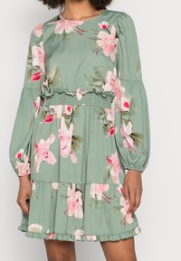 Robe florale en tissu vert doux avec manches longues, taille froncée, ourlet à volants et grandes fleurs roses tout au long du motif.
