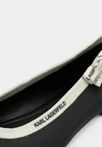 Sapato plano preto com exterior em borracha texturada, contrastado por uma faixa elástica branca com o texto "KARL LAGERFELD." Interior liso.