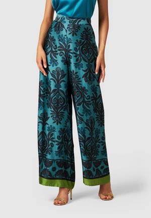 Woman wearing teal wide-leg pants with black floral patterns and green hems, paired with gold high-heeled sandals and a blue top.