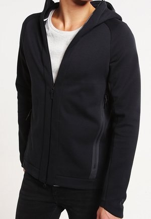 Mężczyzna ubrany w czarną bluzę z zamkiem na białą koszulkę, stojący z rękami swobodnie opuszczonymi wzdłuż ciała na neutralnym tle.