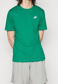 Muž oblečený v zeleném tričku Nike a světlých šortkách, stojící s rukama za zády na jednobarevném pozadí.