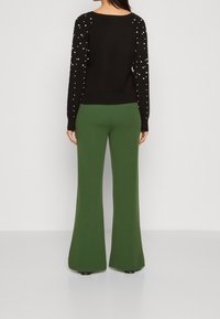 Schwarzer Pullover mit Perlenverzierung an den Ärmeln, kombiniert mit grünen Schlaghosen aus einem glatten, elastischen Stoff.