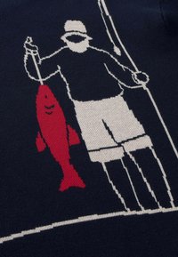 Homme portant un chapeau et un short tenant une canne à pêche et un grand poisson rouge sur un fond en tissu sombre.