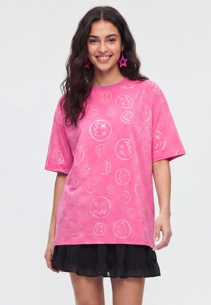 Ung kvinde med langt mørkt hår smiler iført en løs lyserød T-shirt med abstrakte ansigter og et sort flæset skørt.