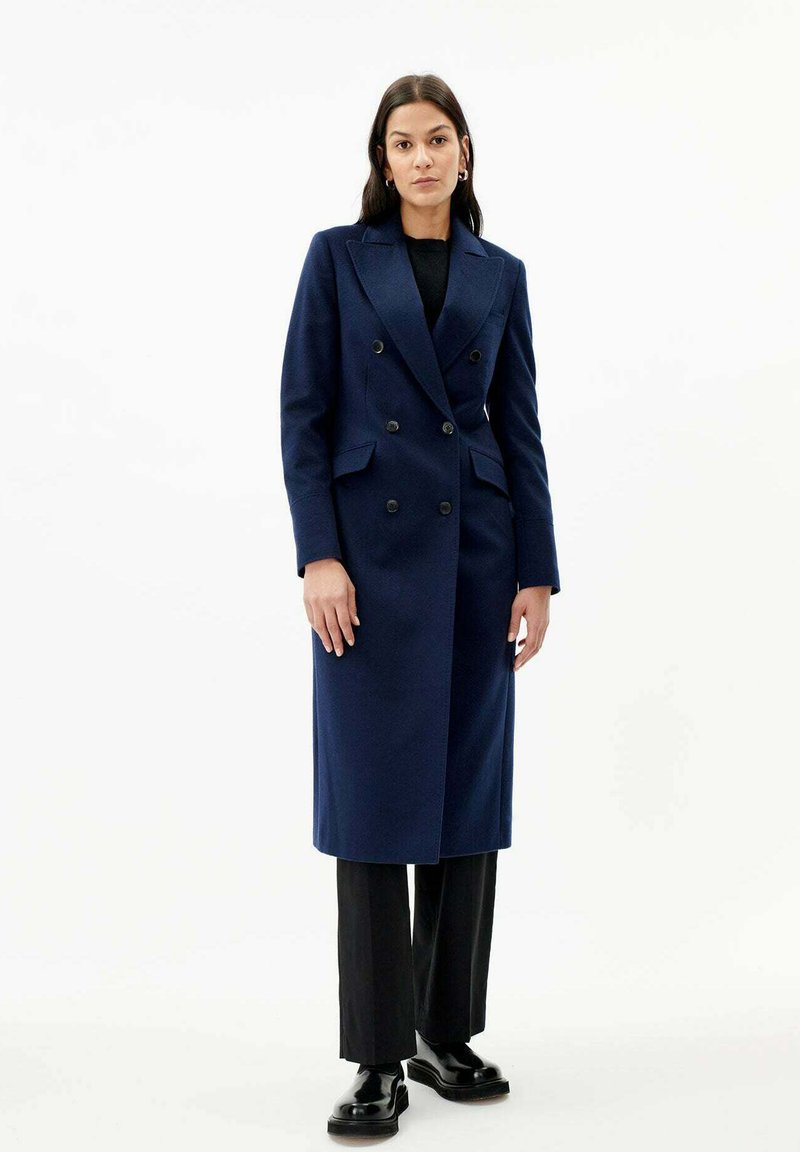 Marinblå dubbelknäppt ullkappa med slag, sidofickor och svarta knappar, i kombination med vida svarta byxor och svarta skor.