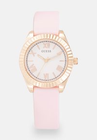 Montre Guess avec un cadran blanc, des chiffres romains dorés et un bracelet en silicone rose. Elle possède une lunette striée dorée et une couronne.