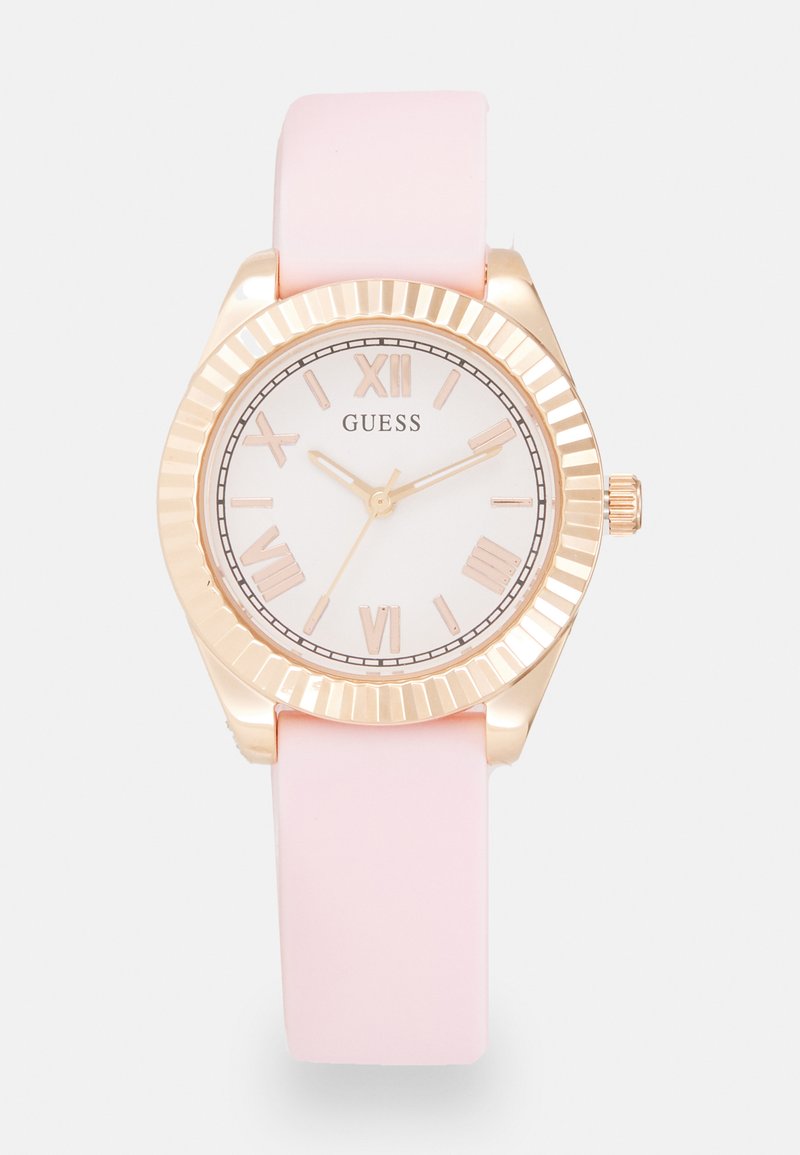 Montre Guess avec un cadran blanc, des chiffres romains dorés et un bracelet en silicone rose. Elle possède une lunette striée dorée et une couronne.