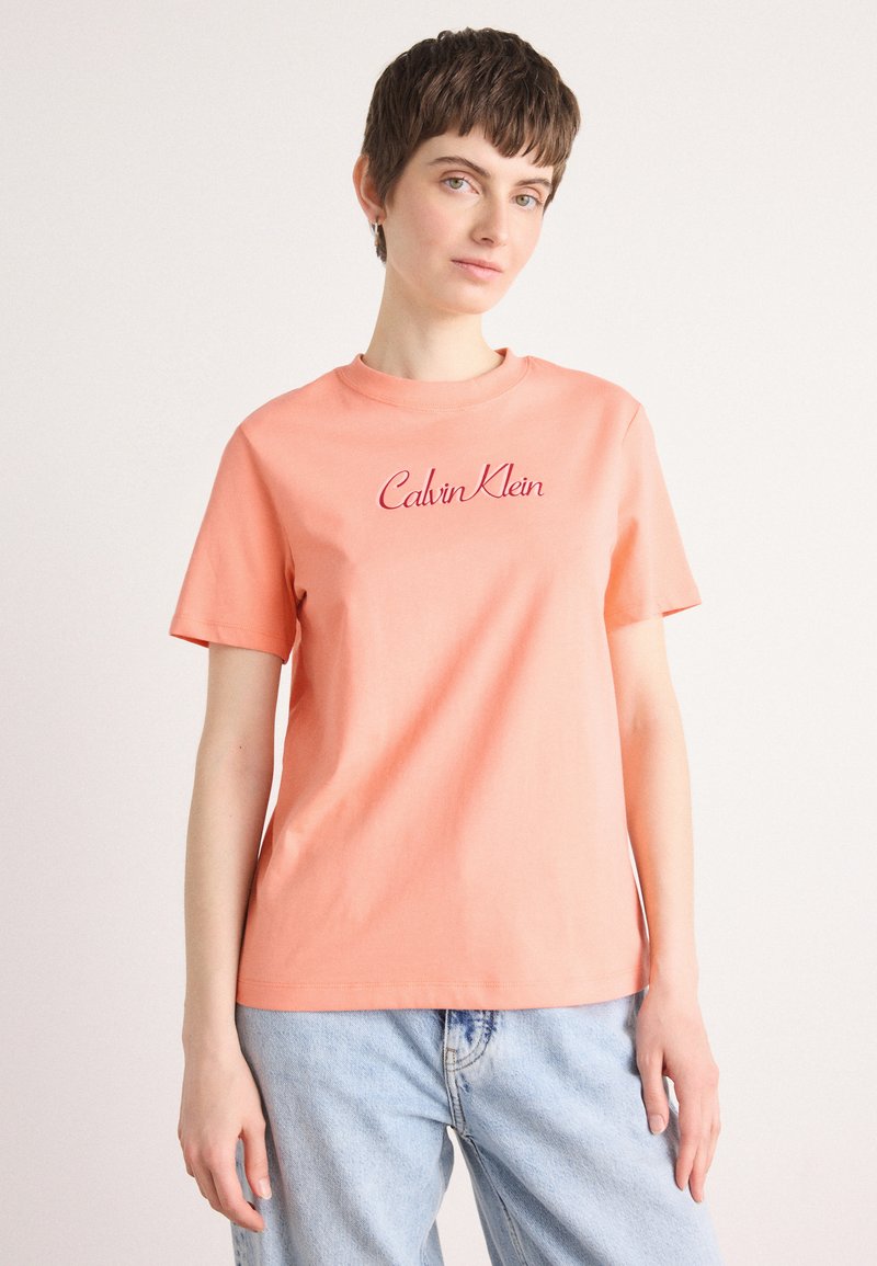 Persikas medvilninis marškinėliai su trumpomis rankovėmis, su raudona siuvinėta "Calvin Klein" logotipu ant krūtinės, apvaliu iškirpimu ir laisvu siluetu.