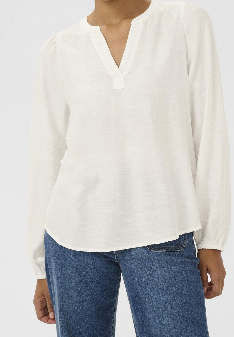 Femme portant un chemisier blanc à manches longues avec un col fendu et un jean en denim bleu, debout avec une main détendue le long du corps.