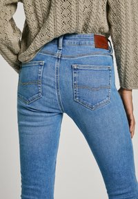 Vaqueros de mezclilla azul claro con bolsillos traseros, usados por una persona con un suéter beige de punto, mostrados desde la cintura hacia abajo sobre un fondo liso.