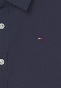 Marineblå knapp-up skjorte med en teksturert stoff. Har sølvknapper og en liten brodert logo i rød, hvit og blå på brystet.