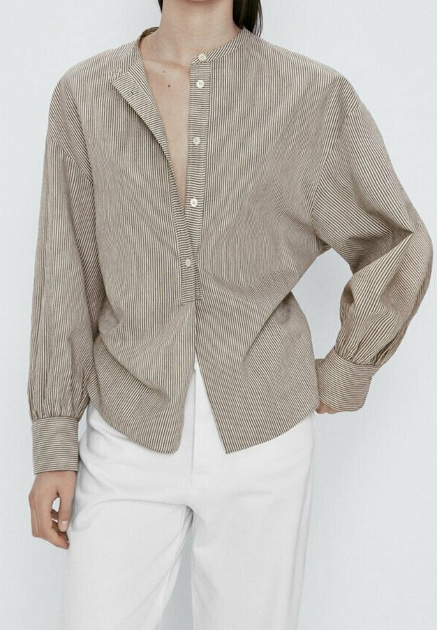 Femme portant une chemise boutonnée rayée verticale beige et blanche à manches longues et un pantalon blanc, debout avec une main sur la hanche.