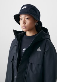 Black bucket hat with white Nike logo, paired with a black jacket featuring pockets and white triangular accents, worn over a black sweatshirt.