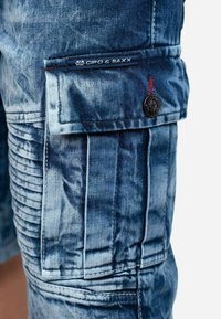 Pantaloni cargo in denim di colore azzurro chiaro con pannelli laterali testurizzati, dettagli con bottoni e un'etichetta cucita sulla vita.