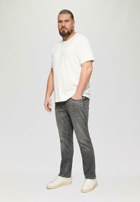 Biała koszulka z krótkim rękawem, szare dżinsy o wyblakłym wyglądzie oraz białe trampki. Model stoi w neutralnej pozycji na jasnym tle.