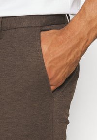 Bruna vävda byxor med sidoficka och synlig sömnad. En hand är placerad i fickan, vilket visar tygets struktur.