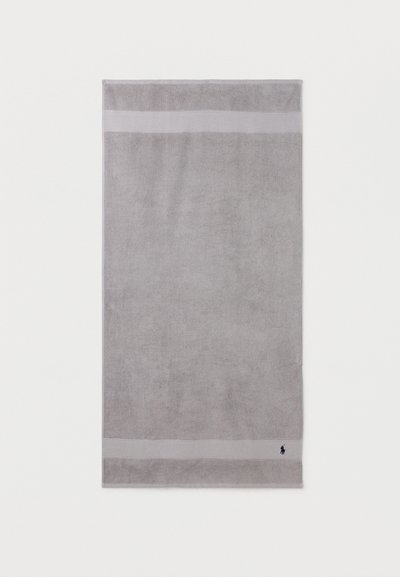 Ανοιχτό γκρι ορθογώνια πετσέτα με δύο οριζόντιες ταινίες κοντά στις άκρες και μικρό σκούρο λογότυπο στην κάτω δεξιά γωνία.