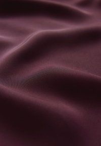 Bordeauxroter Seidenstoff mit einer glatten, glänzenden Oberfläche, der sanfte Falten und eine weiche Textur zeigt und die satte Farbe und den Glanz hervorhebt.