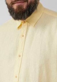 Lichtgele linnen blouse met een col, knopen aan de voorkant en een gestructureerde stof. Close-up weergave met de focus op de details van de blouse.