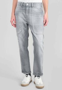 Hombre vestido con jeans gris claro con grandes bolsillos frontales y zapatillas blancas, de pie contra un fondo gris liso.