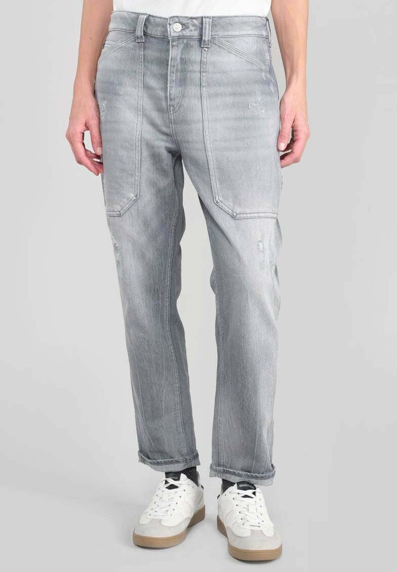 Hombre vestido con jeans gris claro con grandes bolsillos frontales y zapatillas blancas, de pie contra un fondo gris liso.