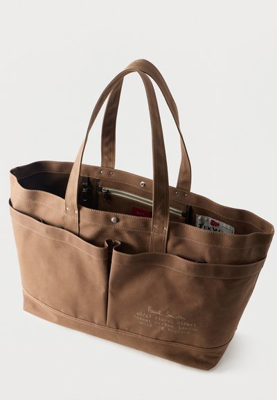 Sac fourre-tout en toile marron avec doubles poignées, poches extérieures à l'avant, poche intérieure zippée, et texte imprimé détaillant une adresse sur le devant.